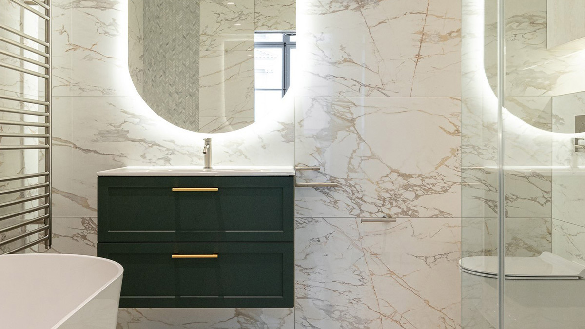 Green and gold bathroom vanity with marble tiles and illuminated mirror