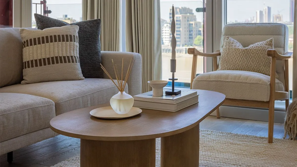 Close-up of a modern living room featuring a wooden coffee table, neutral sofa, accent chair, and floor-to-ceiling windows with riverside views.