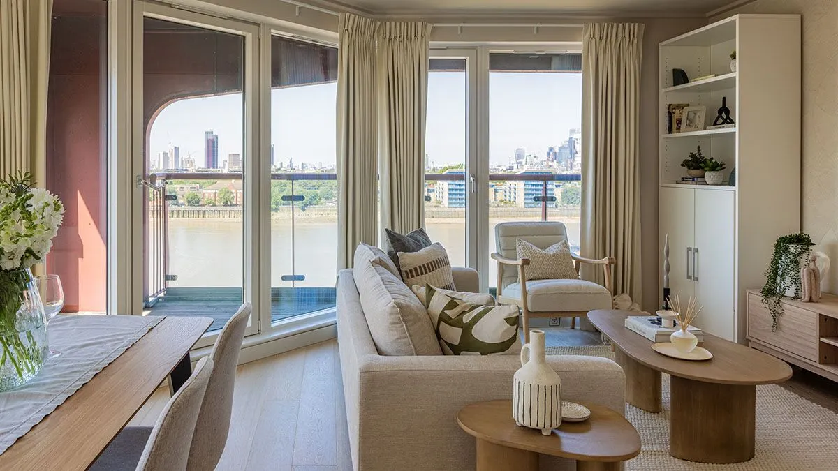 Modern riverside living room with neutral furnishings, floor-to-ceiling windows, balcony access, and panoramic city skyline views.